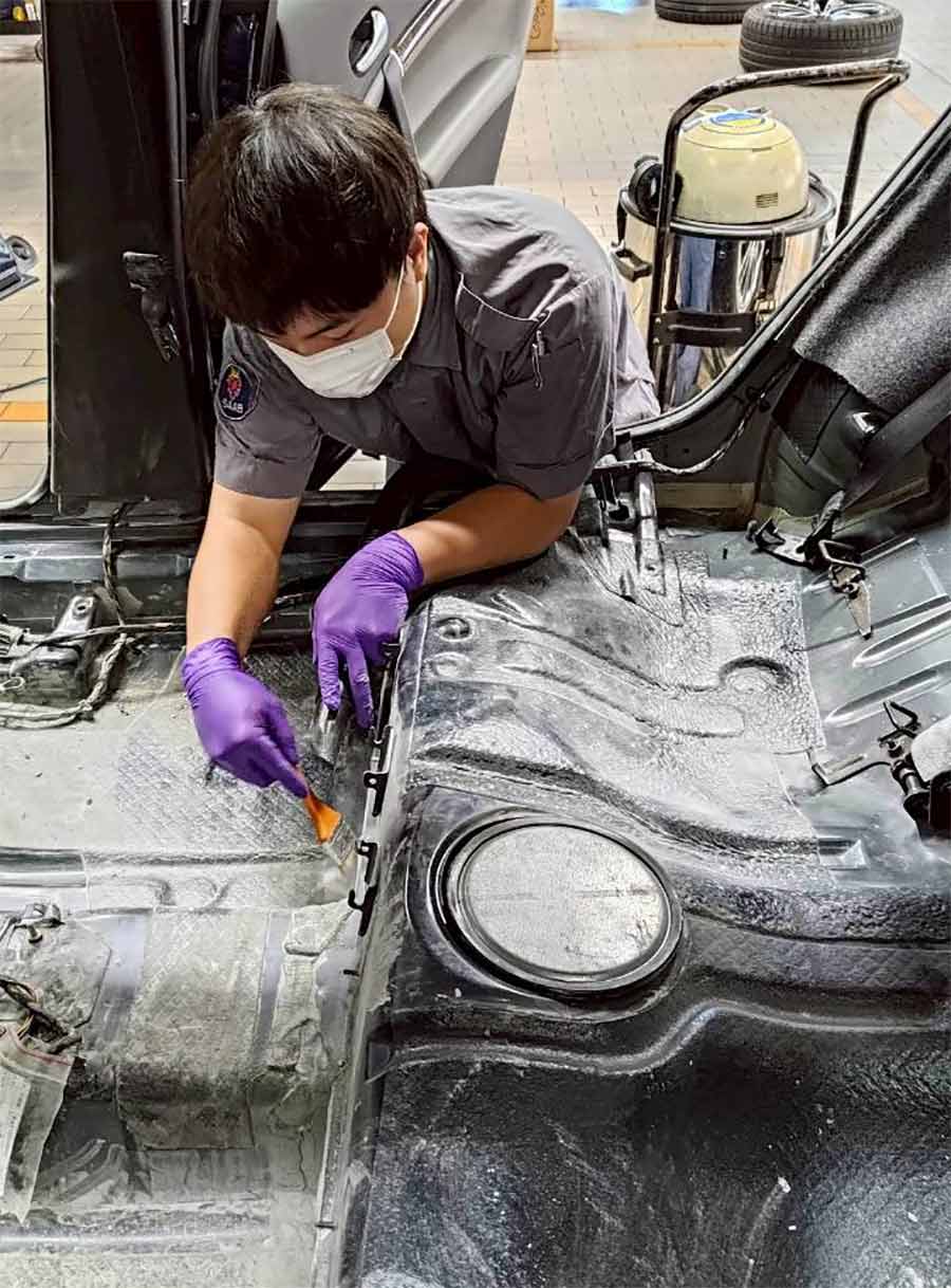 A SAC technician scraping down the exposed floorpan of the Saab 9-5, removing flood residue to bare metal before deep cleaning begins.