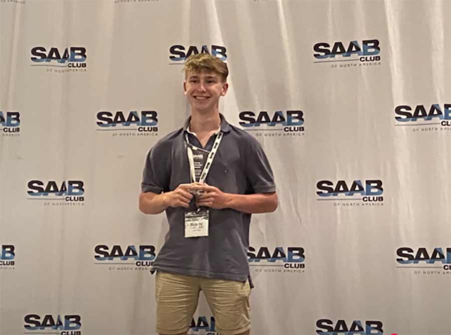 17-year-old Saab enthusiast holding award at Saab Owners Convention 2025, standing in front of Saab Club of North America backdrop.