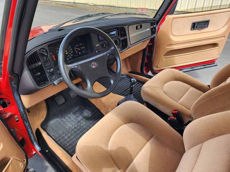 The interior of the 1985 SAAB 900S Coupe invites you into a bygone era, featuring an immaculate tan cloth upholstery, an unblemished dashboard, and a classic manual transmission, all maintained in exceptional condition.