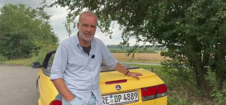 German automotive enthusiast standing beside a yellow Saab 9-3 Convertible during a summer review