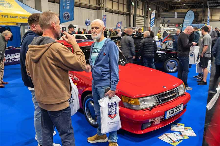 Visitors gathered around a red Saab 900 Carlsson on the Saab Owners Club of Great Britain stand at NEC Classic Motor Show 2025